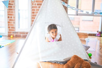 Adorable toddler sitting on the floor inside tipi around lots of toys at kindergarten