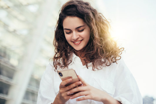 Positive attractive young brunette woman stand outside and look at phone in hands. White blouse and curly hair. Buildings behind. Sun shines and beautiful daylight.