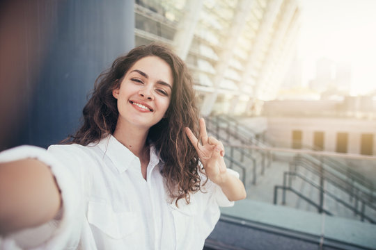 Positive attractive young woman taking selfie in front of building and steps outside. Holding two fingers up. Alone on street. Sunny day. Lovely colors.