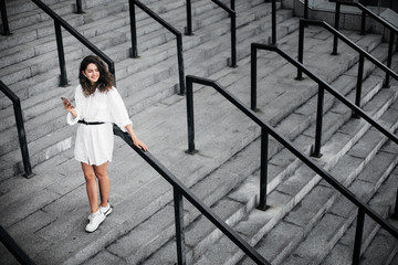 Cheerful modern young woman in white dress stand on grey steps alone and smile. Posing on camera. Stand outside. Positive attractive model hold phone in hand.