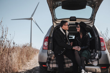 Happy Traveling Couple Enjoying a Car Trip Sitting in Trunk on the Field Road with Electric Wind Turbine Power Generator