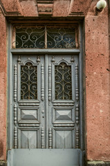 A light blue double wooden door with carved details is covered with openwork wrought iron bars on the windows of the door at the facade of the stone building