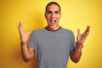 Young handsome man wearing striped t-shirt over yellow isolated background celebrating crazy and amazed for success with arms raised and open eyes screaming excited. Winner concept