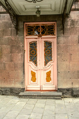 Fototapeta na wymiar Old milky pink double doors with Windows covered with wrought iron bars and figures of Golden color on the facade of a stone house under a canopy in the city of Gyumri