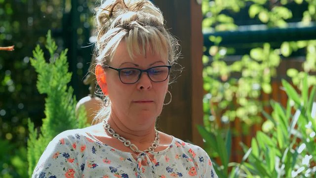 Portrait of 60 years old blond woman with eyeglasses looking at camera at green garden background. 4k slow motion