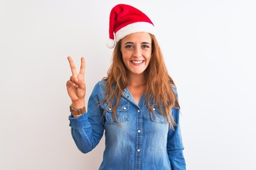 Young beautiful redhead woman wearing christmas hat over isolated background showing and pointing up with fingers number two while smiling confident and happy.