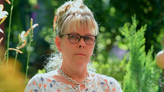 Portrait of 60 years old blond woman with eyeglasses looking at camera at green garden background. 4k slow motion