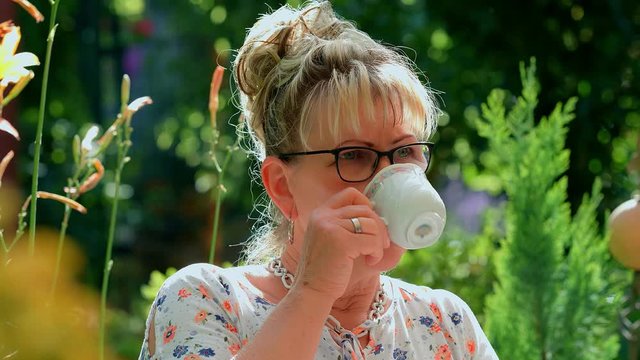 Portrait of 60 years old blond woman with eyeglasses sipping coffee at green garden background. 4k slow motion