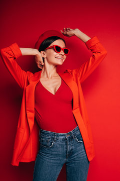 Vertical picture of happy attractive cheerful young woman in red clothes and sunglasses posing on camera. Look to right and simle. Hands up. Isolated over red background.