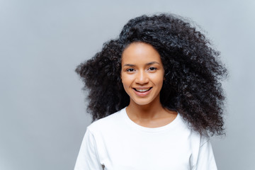 Positive curly woman with natural beauty, dressed in white casual t shirt, has happy expression, looks directly at camera, poses against grey background. Cheerful teenage girl expresses good emotions