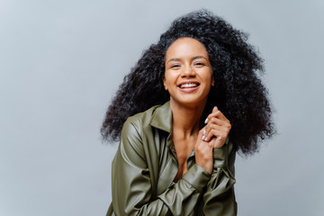 Joyful woman with dark skin, Afro hairstyle, keeps hands together, smiles sincerely at camera,...