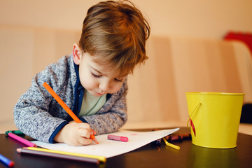 Portrait of small boy two years old sitting on the bed by the table drawing using color pencils and...