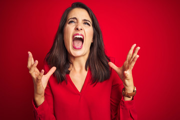 Young beautiful woman wearing shirt standing over red isolated background crazy and mad shouting and yelling with aggressive expression and arms raised. Frustration concept.