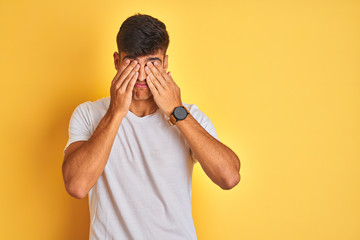 Young indian man wearing white t-shirt standing over isolated yellow background rubbing eyes for...