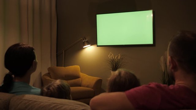 Rear View Of Family With Children Sitting On Sofa In Living Room Evening Watching Green Mock-up Screen TV Together. Family Sitting Together Sofa In Their Living Room Night Watching TV Green Screen.
