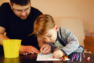 Small boy and his uncle or father sitting by the table at home playing with crayons color pencils drawing and learning family activities having fun