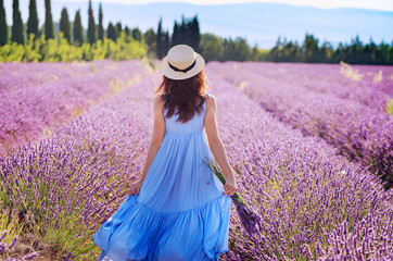Beautiful young woman in boho-chic style dress and hat enjoying the stroll by blooming lavender fields in Provence, France in July. Summer vacations travel background.