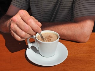 Close-up of a male hand stirring a white cup of cappuccino with a small spoon on a wooden table....