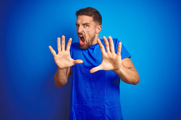 Young handsome man wearing casual t-shirt over blue isolated background afraid and terrified with fear expression stop gesture with hands, shouting in shock. Panic concept.