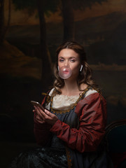 Medieval teen, young girl in vintage dress uses smartphone and blows bubble gum