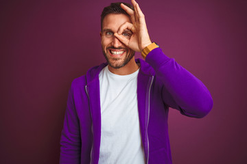 Young fitness man wearing casual sports sweatshirt over purple isolated background doing ok gesture with hand smiling, eye looking through fingers with happy face.