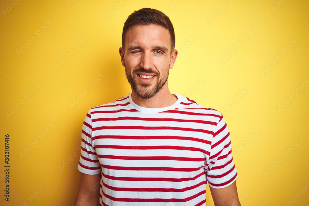 Wall mural Young handsome man wearing casual red striped t-shirt over yellow isolated background winking looking at the camera with sexy expression, cheerful and happy face.