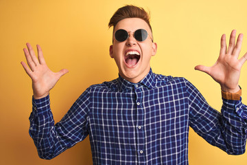 Naklejka premium Young handsome man wearing casual shirt and sunglasses over isolated yellow background celebrating crazy and amazed for success with arms raised and open eyes screaming excited. Winner concept
