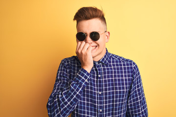 Young handsome man wearing casual shirt and sunglasses over isolated yellow background looking stressed and nervous with hands on mouth biting nails. Anxiety problem.