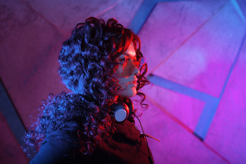 Portrait of young curly pensive sensual woman in big headphones, removed from the head, and shades, on neon background.