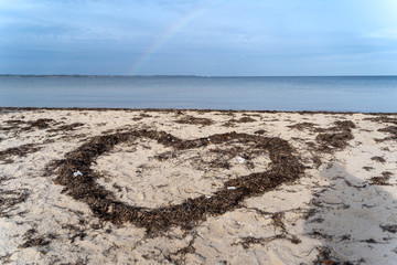 Sandy beach with sea grass in the shape of a heart, rainbow in the background, concept for valentines day and other holidays