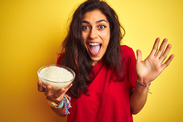 Young beautiful woman holding bowl with rice over isolated yellow background very happy and excited, winner expression celebrating victory screaming with big smile and raised hands