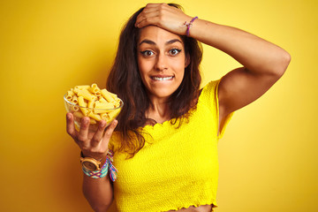 Young beautiful woman holding bowl with dry macaroni pasta over isolated yellow background stressed with hand on head, shocked with shame and surprise face, angry and frustrated. Fear and upset 