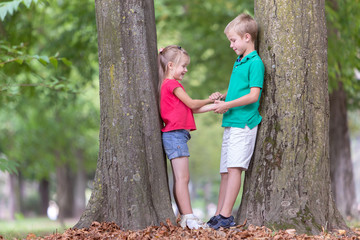 Portrait of two pretty cute children boy and girl standing near big tree trunk in summer park outdoors.