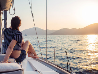 attractive young woman in a striped t-shirt enjoys the sunset on the deck of a sailing yacht....