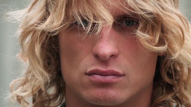 Close up shot of a cool handsome man poprtait. He looks at camera by blue eyes and winds waving his long blond hair