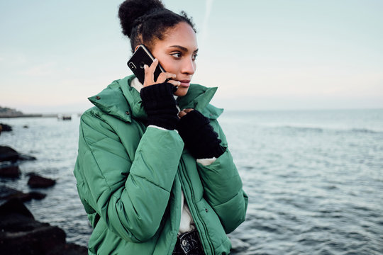 Portrait of attractive African American girl in down jacket talking on cellphone while walking by the sea