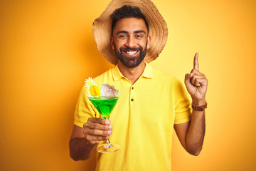 Indian man on vacation drinking cocktail wearing summer hat over isolated yellow background surprised with an idea or question pointing finger with happy face, number one