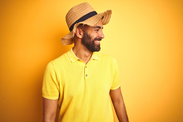 Young indian man on vacation wearing summer hat standing over isolated yellow background looking away to side with smile on face, natural expression. Laughing confident.