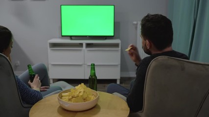 Man and woman are sitting in chairs, watching TV with a green screen, drink beer and eat chips. Back view. Chroma key. Timelapse