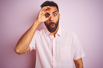 Young indian man wearing casual shirt standing over isolated pink background doing ok gesture shocked with surprised face, eye looking through fingers. Unbelieving expression.