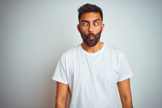 Young indian man wearing t-shirt standing over isolated white background making fish face with lips, crazy and comical gesture. Funny expression.