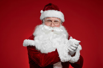 Portrait of Santa Claus in eyeglasses and with white beard showing respect and looking at camera isolated on red background