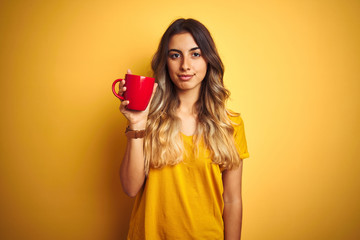 Young beautiful woman holding  red cup of coffee over yellow isolated background with a confident expression on smart face thinking serious