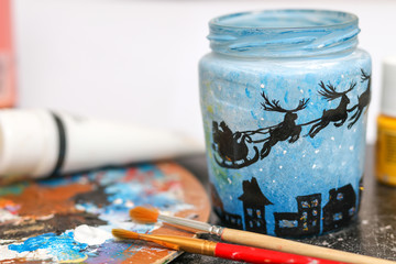 Preparing for the Christmas holidays and New Year.  Drawn on a glass jar Santa on a sleigh with deers.  Artist's supplies, brushes, palette, paints. 