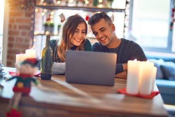 Young beautiful couple sitting using laptop around christmas decoration at home happy face smiling...