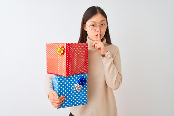 Young chinese woman holding birthday gifts over isolated white background asking to be quiet with finger on lips. Silence and secret concept.