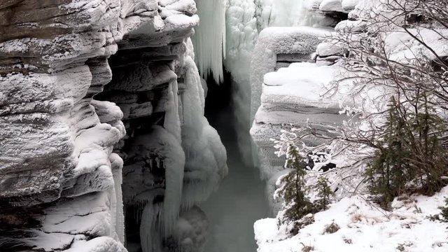 Aerial view of a nearly frozen waterfall in panning right with bare shrub right, Jasper National Park, Alberta Canada