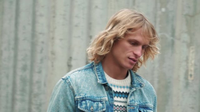 Fashionable young attractive man with blue eyes, long blond curly hair in blue denim jacket walking on urban street on background of the grey wall