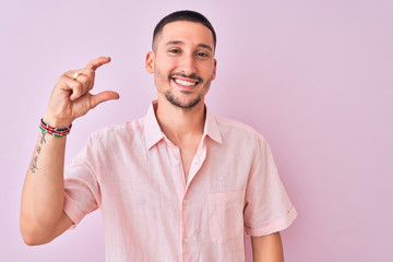 Young handsome man wearing pink shirt standing over isolated background smiling and confident gesturing with hand doing small size sign with fingers looking and the camera. Measure concept.