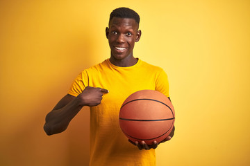 African american athlete man holding basketball ball standing over isolated yellow background with surprise face pointing finger to himself